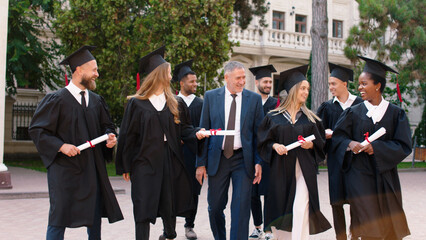 Multiethnic graduates students in the college park walking in group with the college principal after the graduation very exciting holding diplomas and discussing all together
