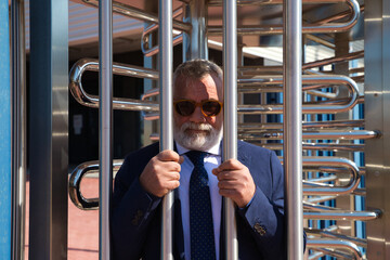 Mature, grey-haired, bearded man with sunglasses behind the bars of a workplace fence. Concept of...