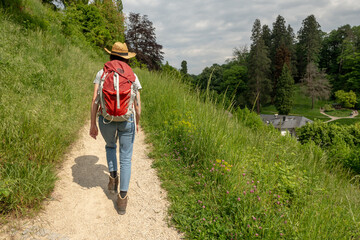 Woman with brown hair, gray t-shirt, jeans, straw hat and red backpack hiking on a trail at...