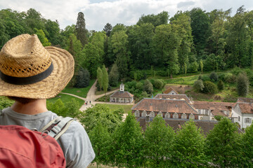 Woman with brown hair, gray t-shirt, straw hat and red backpack looking at Staatspark Fürstenlager Buildings from top of a hill, cloudy sky in the background, rear view