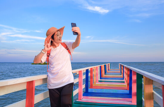 Happy Asian female tourist showing love hand sign while using smartphone to video chat at sea viewpoint against blue sky background