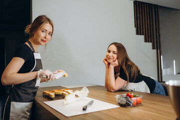 Front view of two confectioners cooking desserts. Beautiful women wearing white and black uniform. They holding confectionery bags and putting dough on baking sheet.