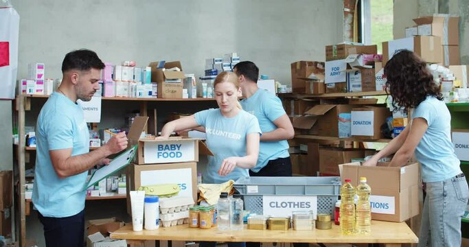 Group of caucasian volunteers in blue uniform taking notes on clipboard and packing donation boxes at warehouse. Young people working at charitable foundation during free time.