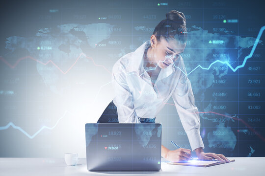 Trading and investing concept with young businesswoman making notes in a book in front of laptop and digital screen with world map, indicators and raising financial chart graph. Double exposure - Powered by Adobe