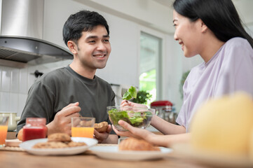 Obraz na płótnie Canvas Young Asian couple enjoying breakfast together. Attractive young Asian woman eating vegetable salad with her husband while having breakfast in kitchen. Healthy food family activity lifestyle concept.