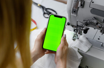 Young beautiful blonde hair seamstress in defocus holding a phone with a green screen on a light background.