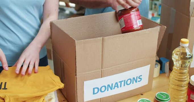 Close up of male and female food bank workers filling cardboard boxes with donated provision at warehouse. Team of caucasian volunteers preparing parcel for delivery to people in need.