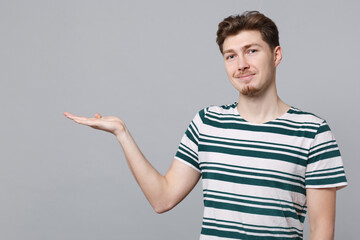 Young smiling happy fun cool caucasian man 20s wearing blue striped t-shirt point hand arm aside on workspace area mock up isolated on plain gray background studio portrait. People lifestyle concept.