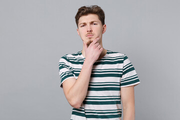 Young puzzled thoughtful confused minded pensive caucasian man 20s wearing blue striped t-shirt look camera prop up chin isolated on plain gray background studio portrait. People lifestyle concept