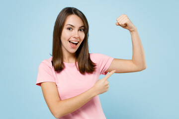 Young happy woman 20s in pink t-shirt showing point index finger on biceps muscles on hand demonstrating strength power isolated on pastel plain light blue background studio. People lifestyle concept.