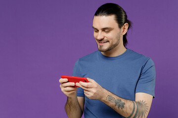 Gambling young smiling fun man 20s in basic blue t-shirt using play racing app on mobile cell phone hold gadget smartphone for pc video games isolated on plain purple color background studio portrait