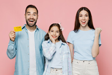 Young happy fun parents mom dad with child kid daughter teen girl in blue clothes hold in hand credit bank card do winner gesture isolated on plain pastel light pink background. Family day concept
