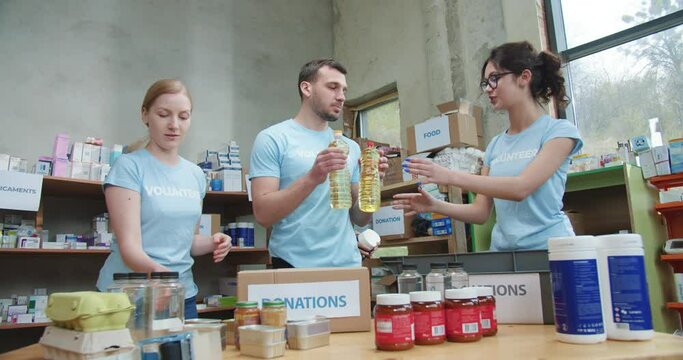 Young Team In Blue T-shirts Packing Food And Donation Clothes To Boxes At Charity Warehouse. Friendly Brigade Of Local Volunteers Sending Humanitarian Aid For Homeless And Needy People.