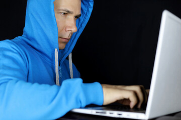 Man in and blue hoodie working at laptop on black background. Concept of cybercrime, hacking or...