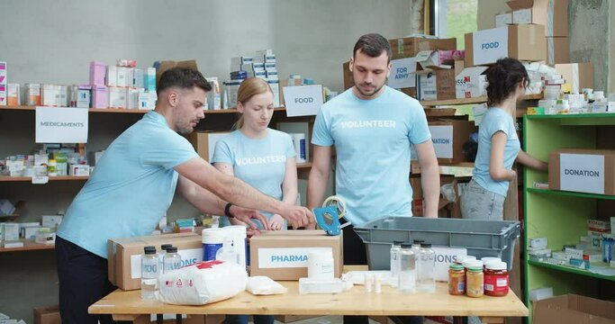 Group of people packing medicines for people in need while working at humanitarian aid project. Caucasian man in blue t-shirt sticking with scotch cardboard donation boxes.