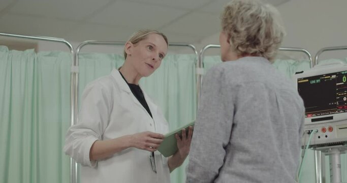 Female Doctor Examining Senior Patient in Hospital, taking notes on Digital Tablet