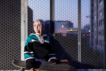 Young blonde hair hipster girl with skateboard sitting on bench in the city at night.
