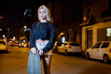 Fototapeta premium Front view of young pretty woman looking camera in shorts while standing on the street holding a skateboard at night in the city