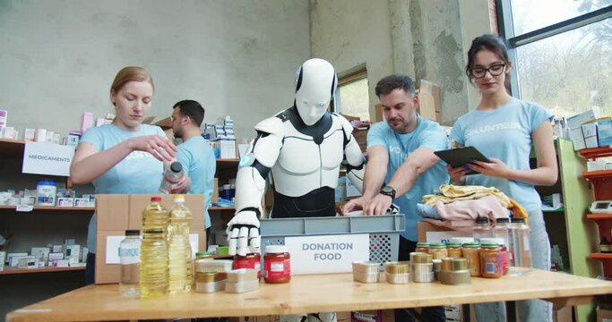 Aid workers with android robot putting food and drinks into boxes for delivery to people in need. Volunteers and robotic cyborg working together at charity warehouse and using digital tablet.