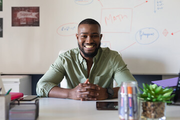 Portrait of smiling african american young male teacher sitting at desk against whiteboard in class
