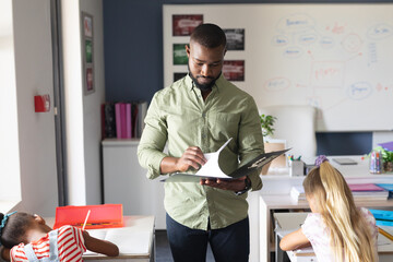 African american young male teacher reading file while standing by multiracial girls in classroom - Powered by Adobe