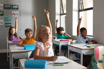Multiracial elementary school students raising hands while sitting at desk in classroom