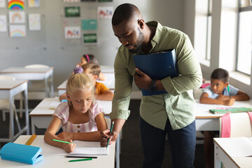African american young male teacher assisting caucasian elementary schoolgirl at desk in classroom - obrazy, fototapety, plakaty