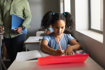African american elementary schoolgirl writing on book at desk in classroom