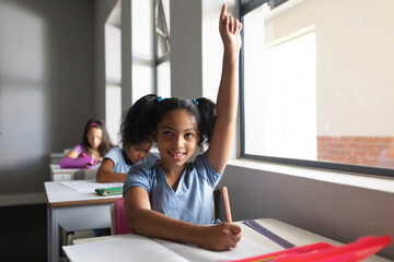 Smiling biracial elementary school girl with hand raised sitting at desk in classroom - obrazy, fototapety, plakaty