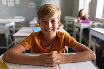 Portrait of smiling caucasian elementary schoolboy with hands clasped sitting at desk in classroom