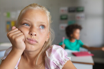 Close-up of caucasian elementary schoolgirl with hand on chin looking away while sitting at desk