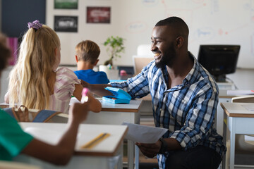 Smiling african american young male teacher talking to caucasian elementary schoolgirl at desk