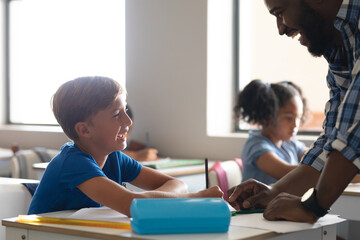 Smiling african american young male teacher talking to caucasian elementary schoolboy at desk