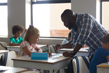 Fototapeta premium African american young male teacher teaching to caucasian elementary schoolgirl sitting at desk