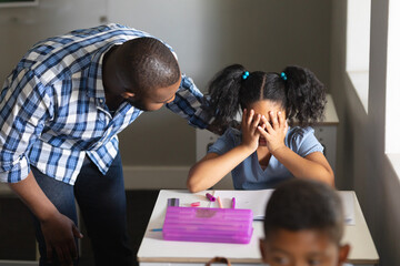 African american young male teacher consoling sad biracial elementary schoolgirl sitting at desk