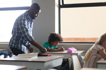 African american young male teacher assisting african american elementary schoolboy studying at desk