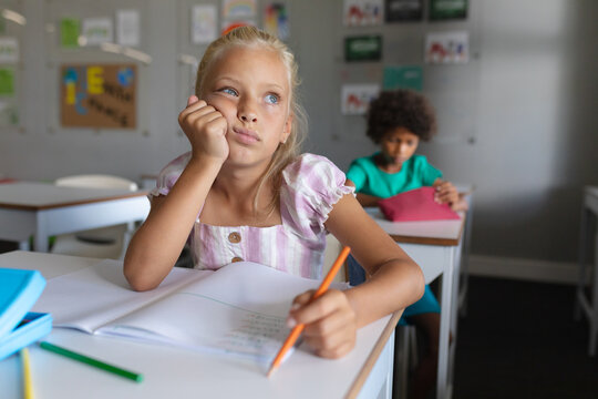 Caucasian elementary schoolgirl with hand on chin looking away while studying at desk in classroom