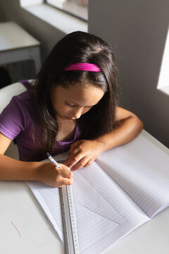 High angle view of caucasian elementary schoolgirl drawing line on book at desk in classroom