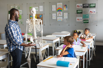 Smiling african american young male teacher explaining skeleton to multiracial elementary students