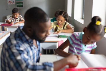 Fototapeta premium African american young male teacher assisting african american elementary girl studying at desk