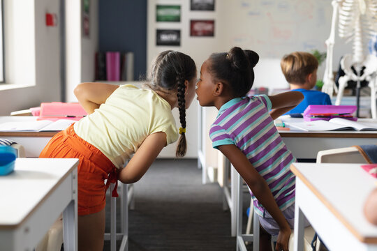 African american elementary girl whispering to caucasian female classmate at desk in classroom