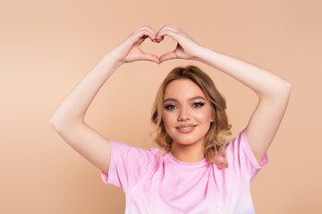 Fototapeta premium happy woman in t-shirt showing heart sign above head isolated on beige.