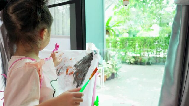 Close up of painting process of Asian child girl standing and hold paint brush drawing black color to canvas on an easel in playroom.Try to use paint brush with black color. Children's creativity. h