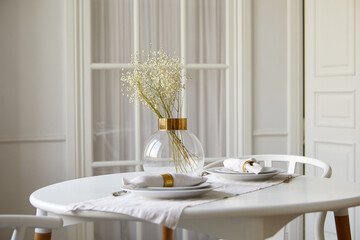 Elegant glass vase with gentle babys breath flowers and ceramic plates with rolled napkins served on round table in white dining room