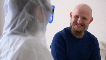 The doctor came to the patient and amuses him in order to cheer up the patient. A doctor in a mask and protective suit is fooling around with a patient, a middle-aged man with a beard.
