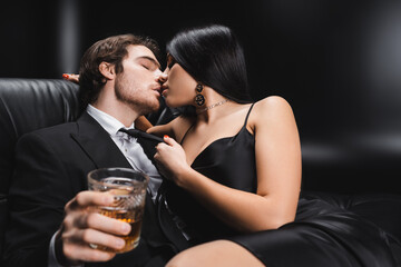 Sensual brunette woman kissing boyfriend in suit holding blurred whiskey on black background.