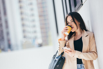 Beautiful confident female entrepreneur dressed in stylish suit drinking coffee and using mobile phone while standing on street against wall