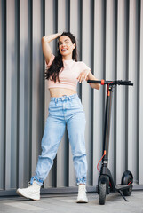A young beautiful hipster woman is standing next to her electric scooter with modern architecture in the background.