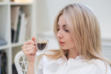 A young beautiful Caucasian blonde woman rests at home. A woman sits at a table, drinking coffee from a glass cup and reading a magazine.