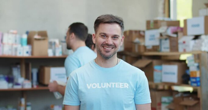 Portrait of bearded caucasian man in blue volunteer t-shirt smiling sincerely on camera while standing at warehouse of food bank. Donation and charity concept.